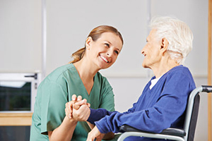 Photo of a woman in green scrubs holding the hand of an elderly woman in a wheelchair, both are smiling